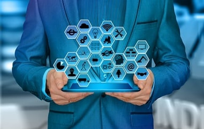 Użytkownik trzyma tablet, na którym wyświetlane są ikony w hexagonalnych kształtach. Ikony przedstawiają różne usługi i technologie, takie jak komunikacja, transport i nauka. Tło jest rozmyte, z odcieniami niebieskiego.